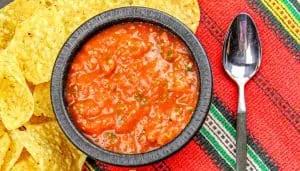 Bowl of red salsa surrounded by tortilla chips on a colorful red, green, and yellow striped cloth with a spoon beside it.
