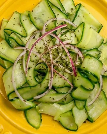 Cucumber ribbons and thin red onion slices seasoned with herbs on a bright yellow plate over a beige cloth.
