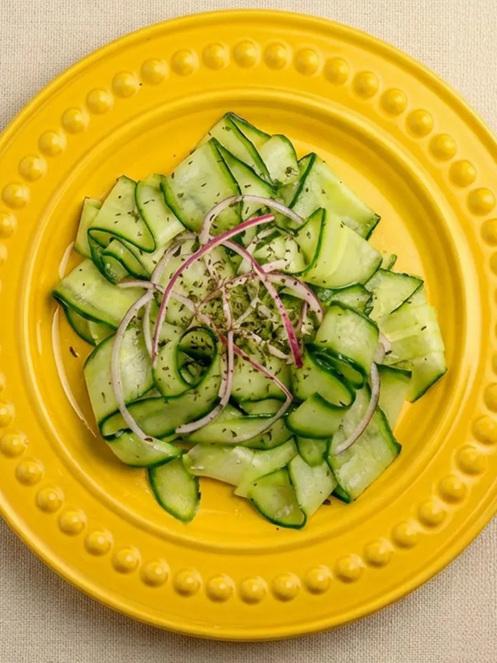 Thin cucumber ribbons and red onion slices seasoned with herbs on a yellow plate with raised dot border, on a beige fabric surface.