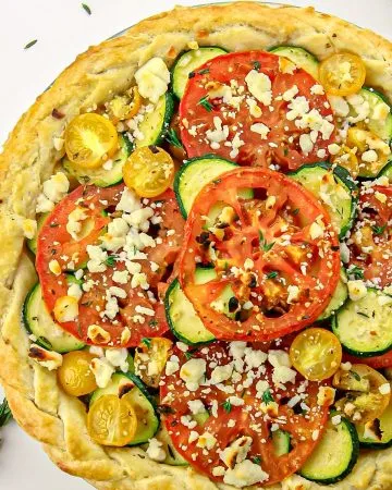 Tomato, zucchini, and yellow cherry tomato tart topped with crumbled cheese and fresh thyme sprigs on a white background.