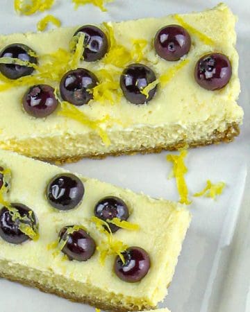 Two rectangular cheesecake slices topped with blueberries and lemon zest on a white plate.