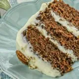 Slice of three-layer carrot cake with cream cheese frosting on blue plate, garnished with pecan half, next to floral napkin.