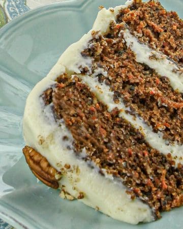 Slice of three-layer carrot cake with cream cheese frosting on blue plate, garnished with pecan half, next to floral napkin.