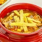 Red bowl of chicken tortilla soup topped with tortilla strips, with a plate of Mexican rice partially visible in the background.