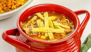 Red bowl of chicken tortilla soup topped with tortilla strips, with a plate of Mexican rice partially visible in the background.