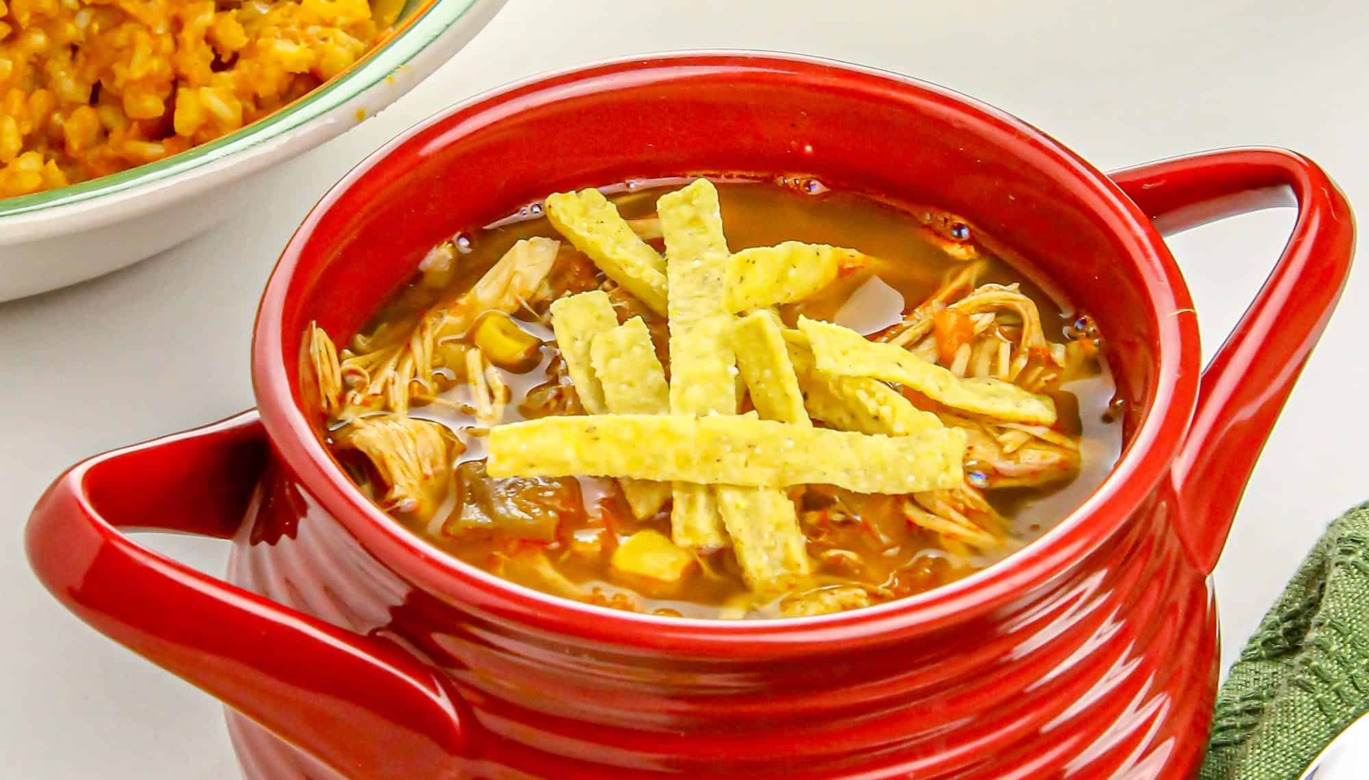 Red bowl of chicken tortilla soup topped with tortilla strips, with a plate of Mexican rice partially visible in the background. Soup recipes