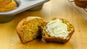 Golden-brown muffin cut in half, one half spread with butter, on a wooden surface with muffin tin and cooling rack nearby.