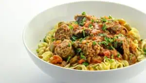 Bowl of egg noodles topped with sausage slices in tomato-based sauce garnished with chopped parsley.