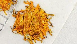 Crispy golden-brown shredded sweet potato latke with black pepper seasoning on paper towel, next to metal cooling rack.