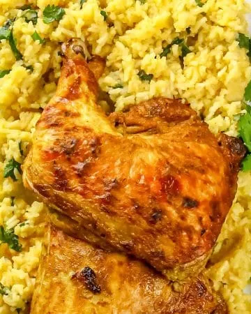 Roasted chicken leg quarters on yellow rice garnished with cilantro on a white rectangular plate with green patterned tablecloth.