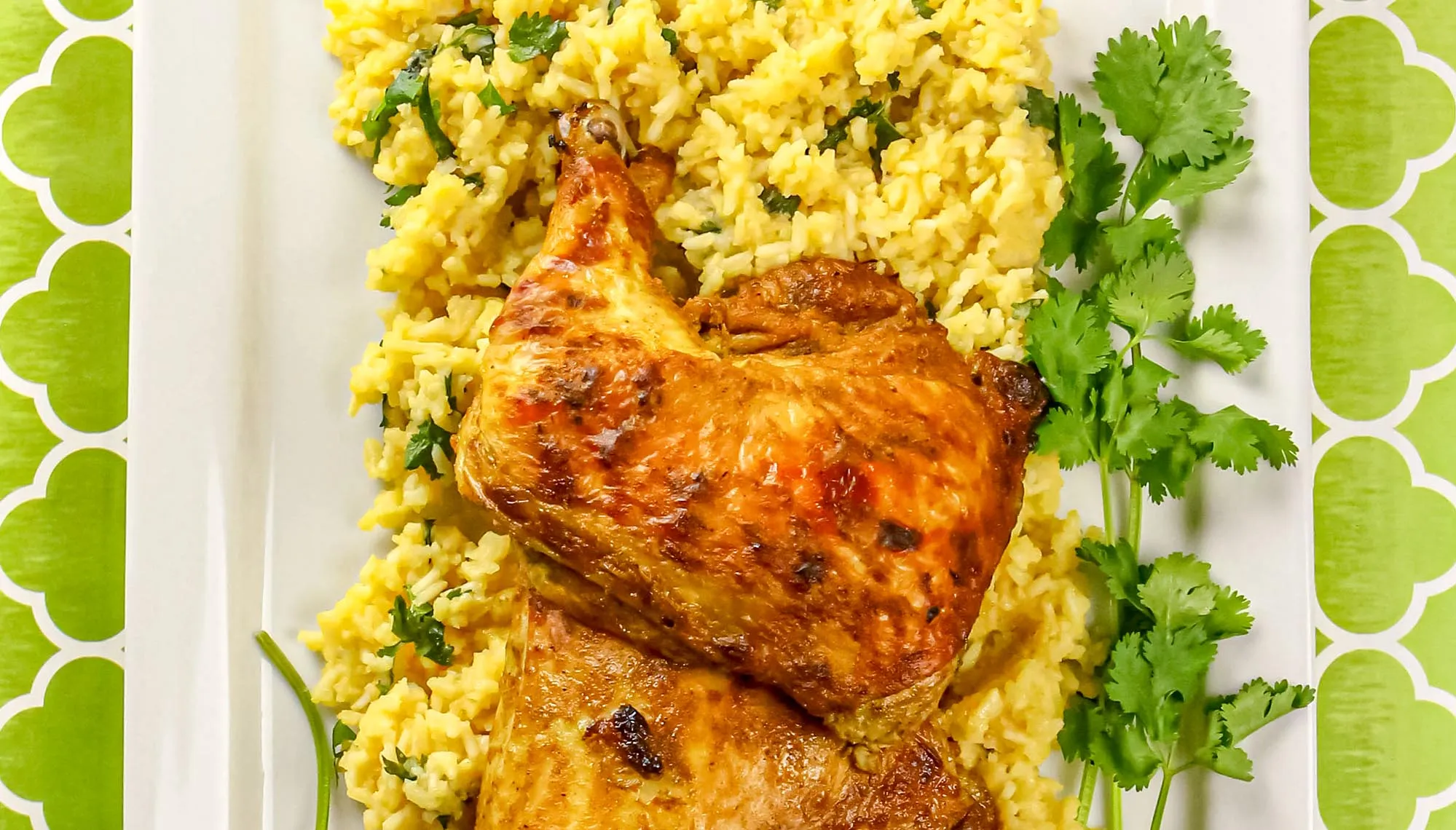 Roasted chicken leg quarters on yellow rice garnished with cilantro on a white rectangular plate with green patterned tablecloth.