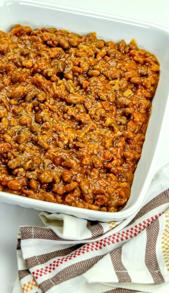Close-up of baked beans in a white square dish on a striped cloth napkin.