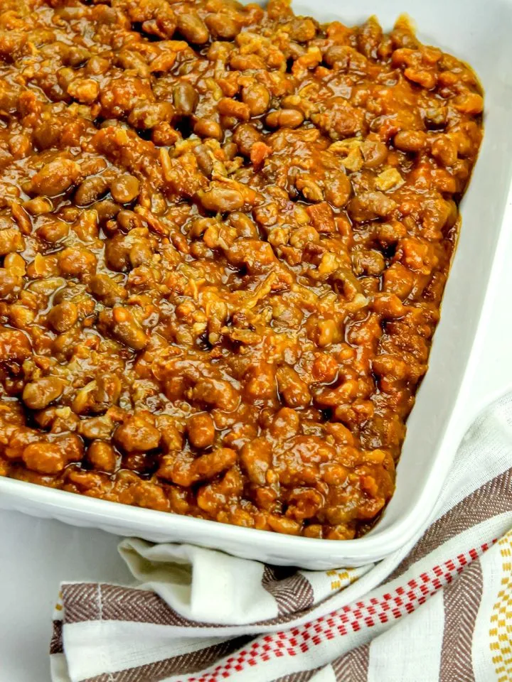 Close-up of baked beans in a white square dish on a striped cloth napkin.