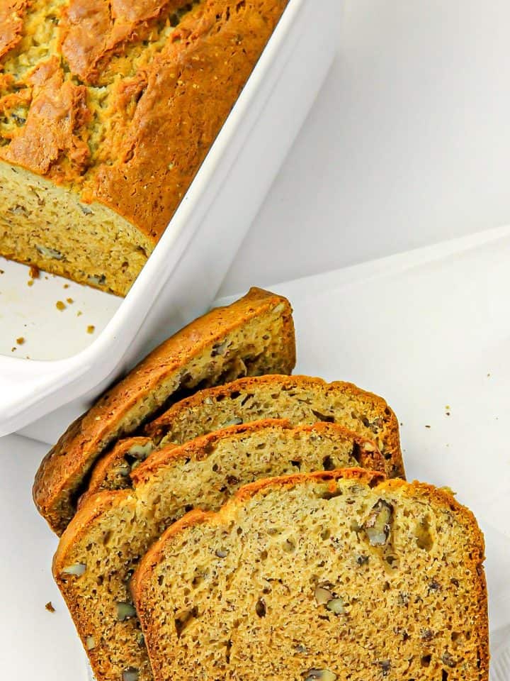 Sliced nut bread with visible walnut pieces on a white surface beside a partially sliced loaf in a white baking dish.