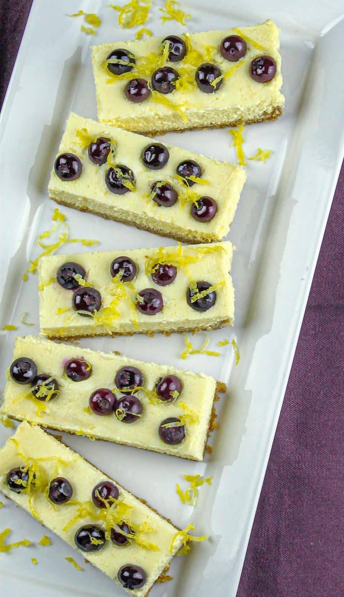 Five rectangular lemon cheesecake bars topped with blueberries and lemon zest on a white platter.