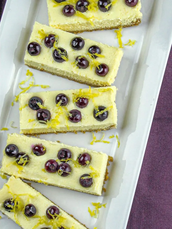 Five rectangular lemon cheesecake bars topped with blueberries and lemon zest on a white platter.