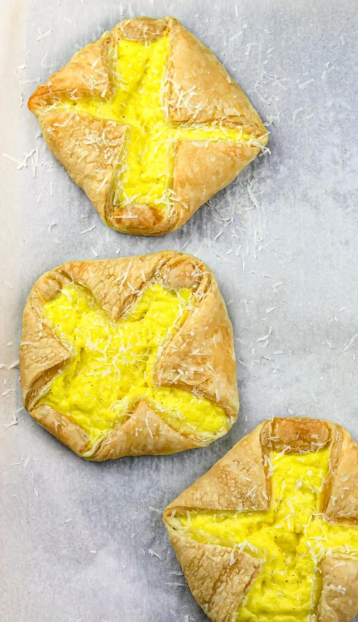 Three puff pastry tarts filled with scrambled eggs, sprinkled with grated cheese on a parchment-lined tray.
