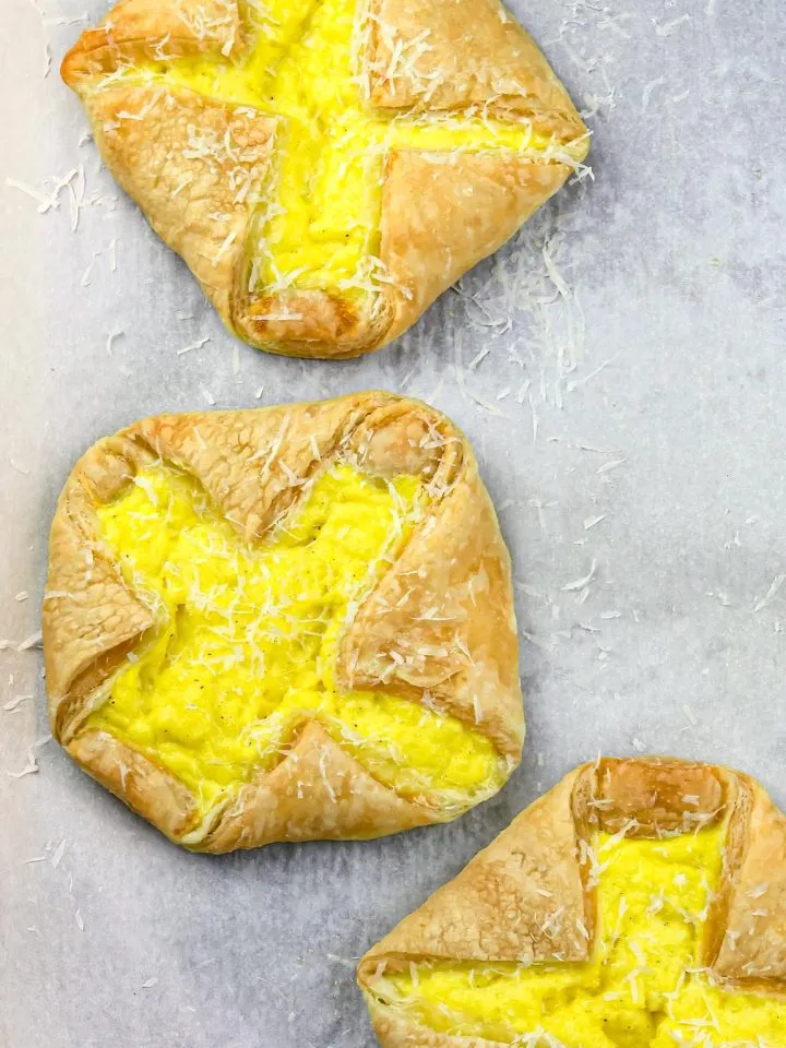 Three puff pastry tarts filled with scrambled eggs, sprinkled with grated cheese on a parchment-lined tray.