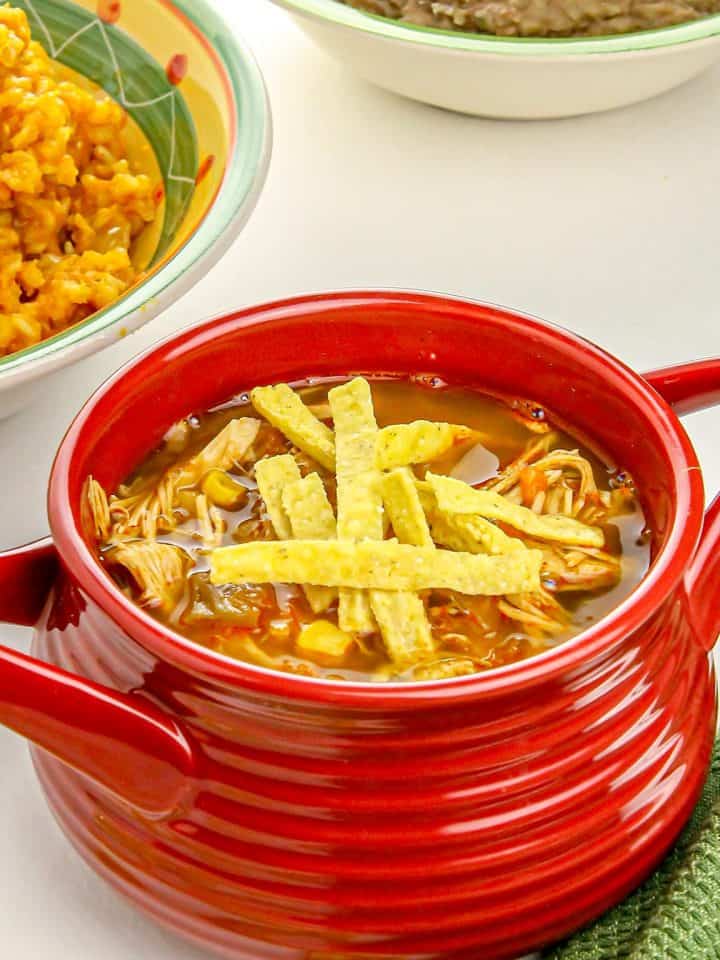 Chicken tortilla soup in a red bowl topped with tortilla strips, with bowls of refried beans and Mexican rice in the background.
