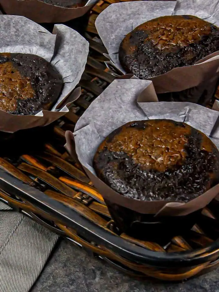Four chocolate muffins in brown paper liners in a wicker basket on a gray surface.