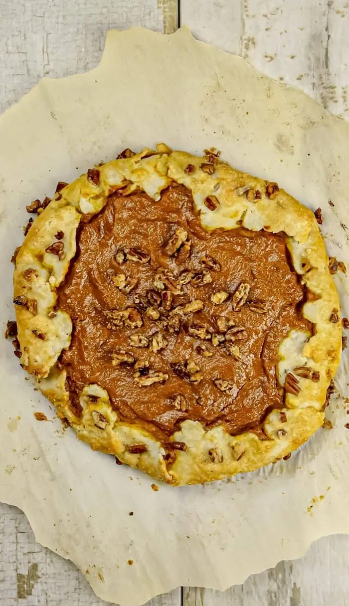 Rustic galette with pumpkin filling topped with chopped pecans on parchment paper over wooden surface.