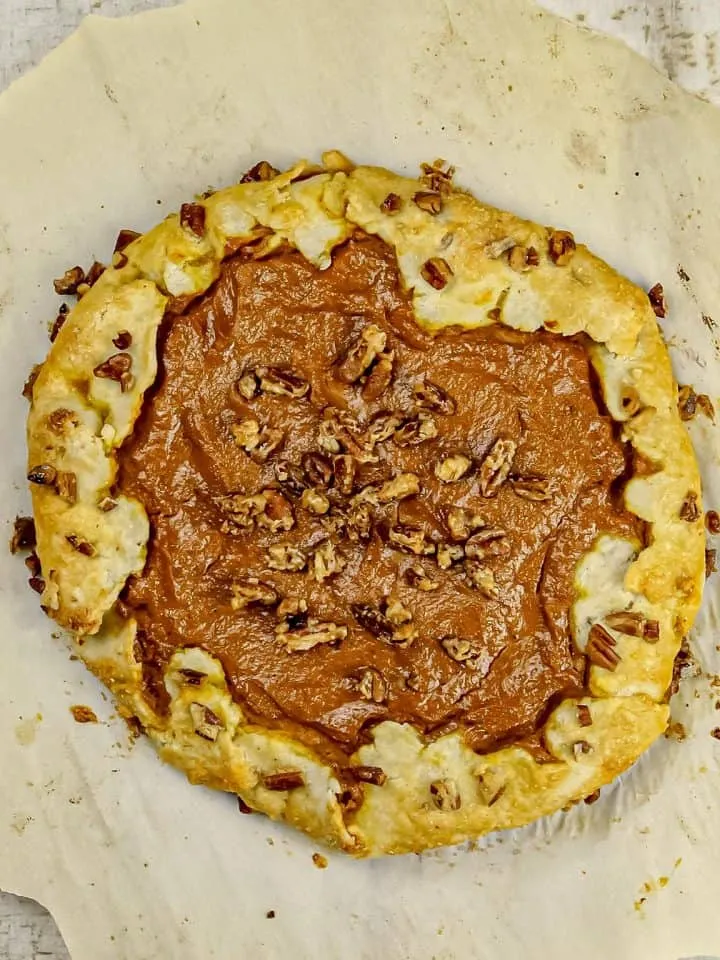 Rustic galette with pumpkin filling topped with chopped pecans on parchment paper over wooden surface.