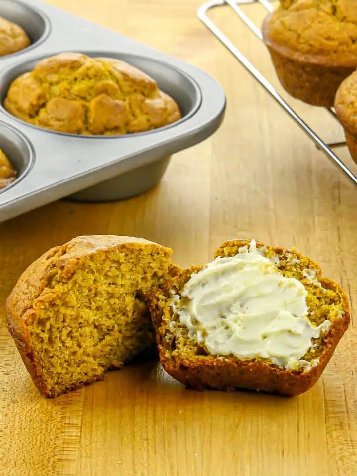Moist split pumpkin muffin with creamy butter spread on one half, resting on a wooden surface, with more muffins in a baking tray and cooling rack behind.