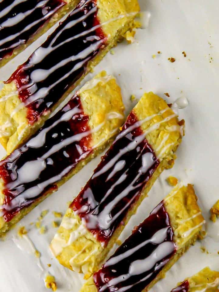 Jam-filled shortbread bars drizzled with white icing on a white surface with crumbs scattered around.