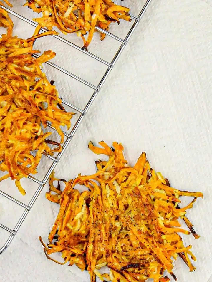 Golden brown, crispy shredded sweet potato latkes on a cooling rack and paper towel, seasoned with black pepper.