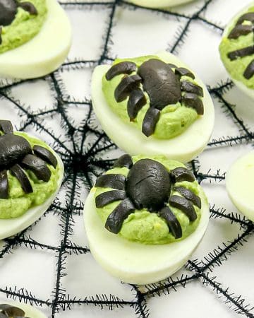 Deviled eggs with green filling topped with black olive pieces arranged to look like spiders, placed on a black spider web decoration.