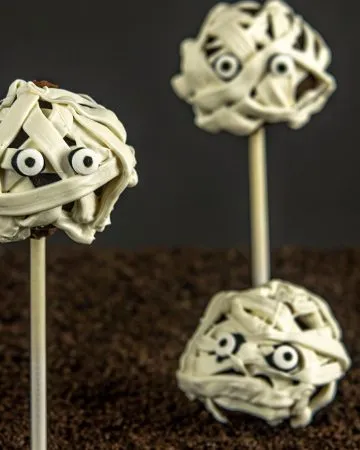 Three mummy-themed cake pops with white icing bandages and candy eyes on sticks, set against a dark background.
