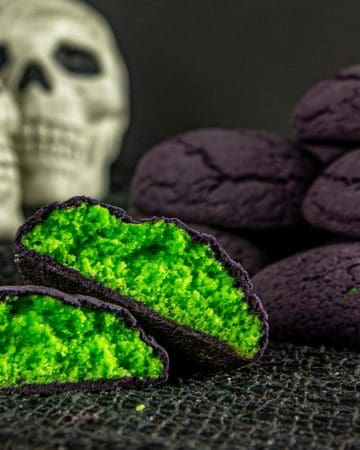 Black cookies with bright green filling, one cookie split in half, stacked cookies in the background, two white skull decorations behind them.