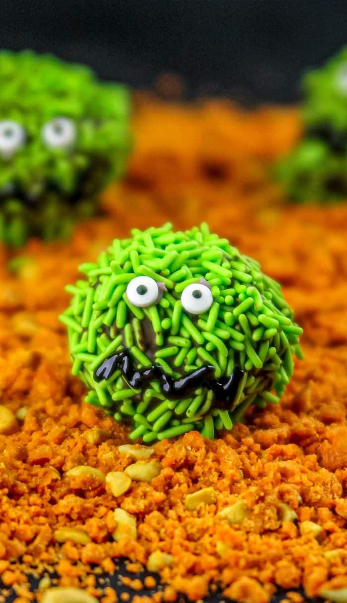 Green sprinkle-covered treats with candy eyes and chocolate mouths on an orange crumbly surface.