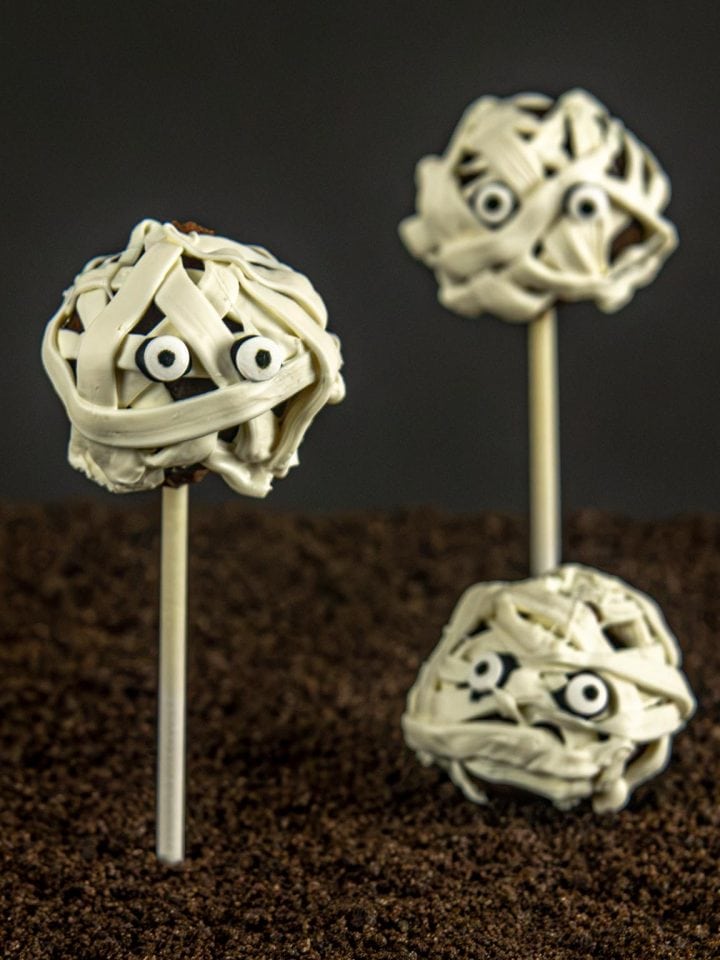 Three mummy-themed cake pops with white chocolate "bandages" and candy eyes, standing in a bed of crushed cookies against a dark background.