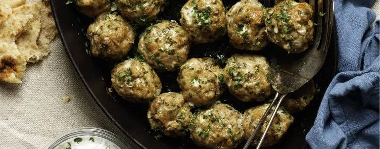 Herbed meatballs in a black pan with flatbread on a beige cloth and a blue napkin nearby.