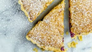 Three raspberry tarts dusted with powdered sugar on a gray surface, one tart is sliced into a triangle.