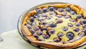 Baked cherry clafoutis with golden crust and dark cherries in a round metal pan on a pale cloth.