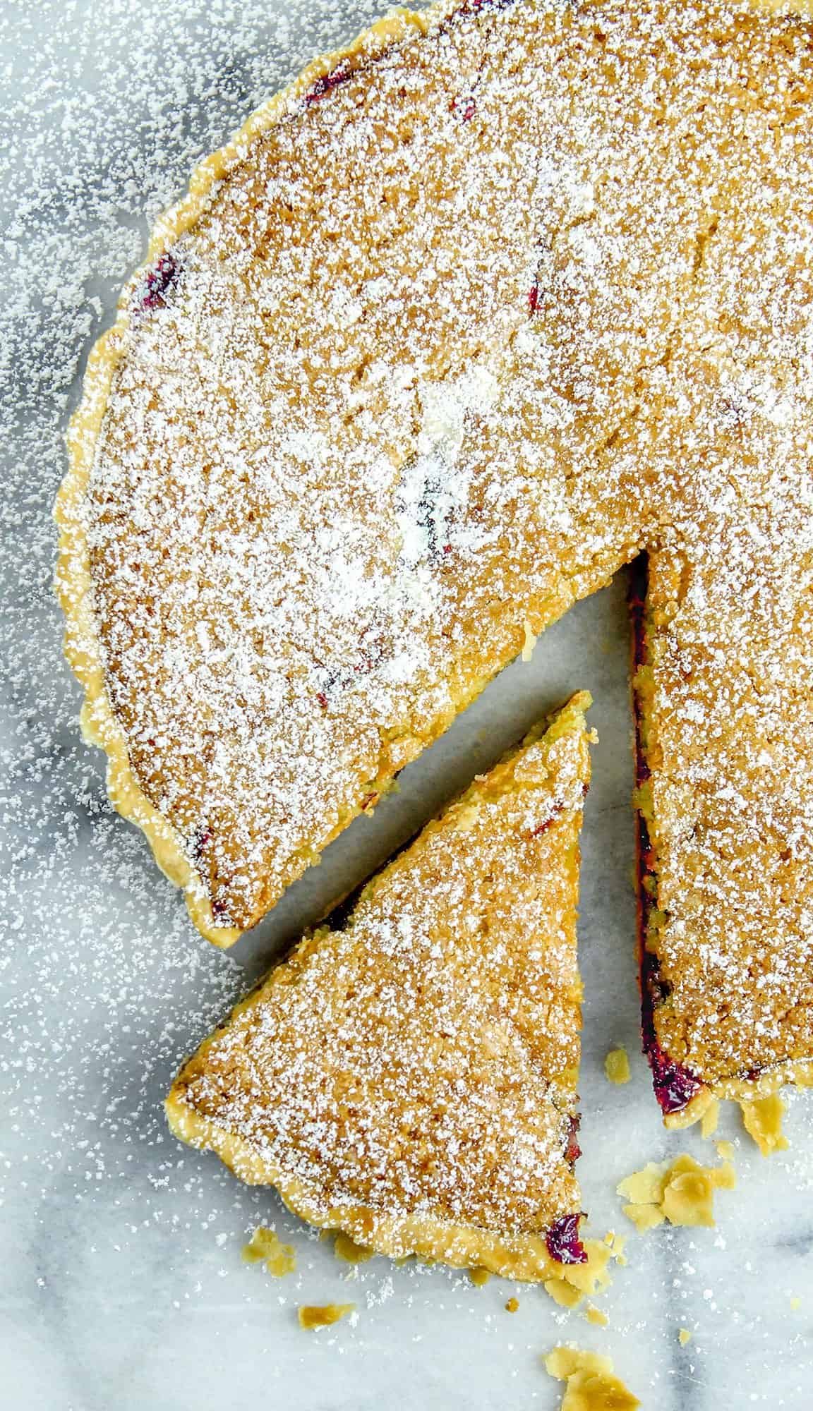 Round tart dusted with powdered sugar, one slice removed and placed nearby on marble surface with scattered crumbs.