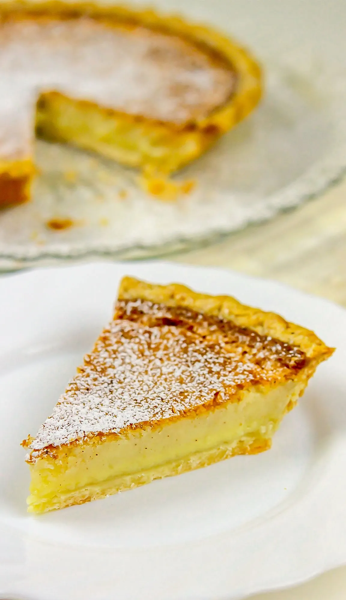 Slice of custard pie dusted with powdered sugar on a white plate, with the remaining pie in the background.