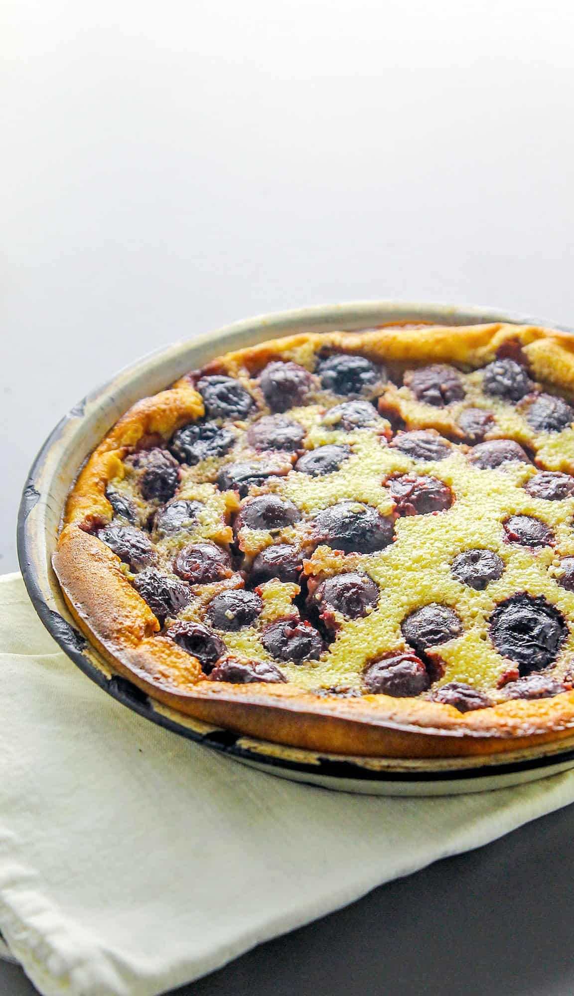 Round cherry clafoutis with golden crust on a baking pan, placed on a folded white cloth on a dark surface.