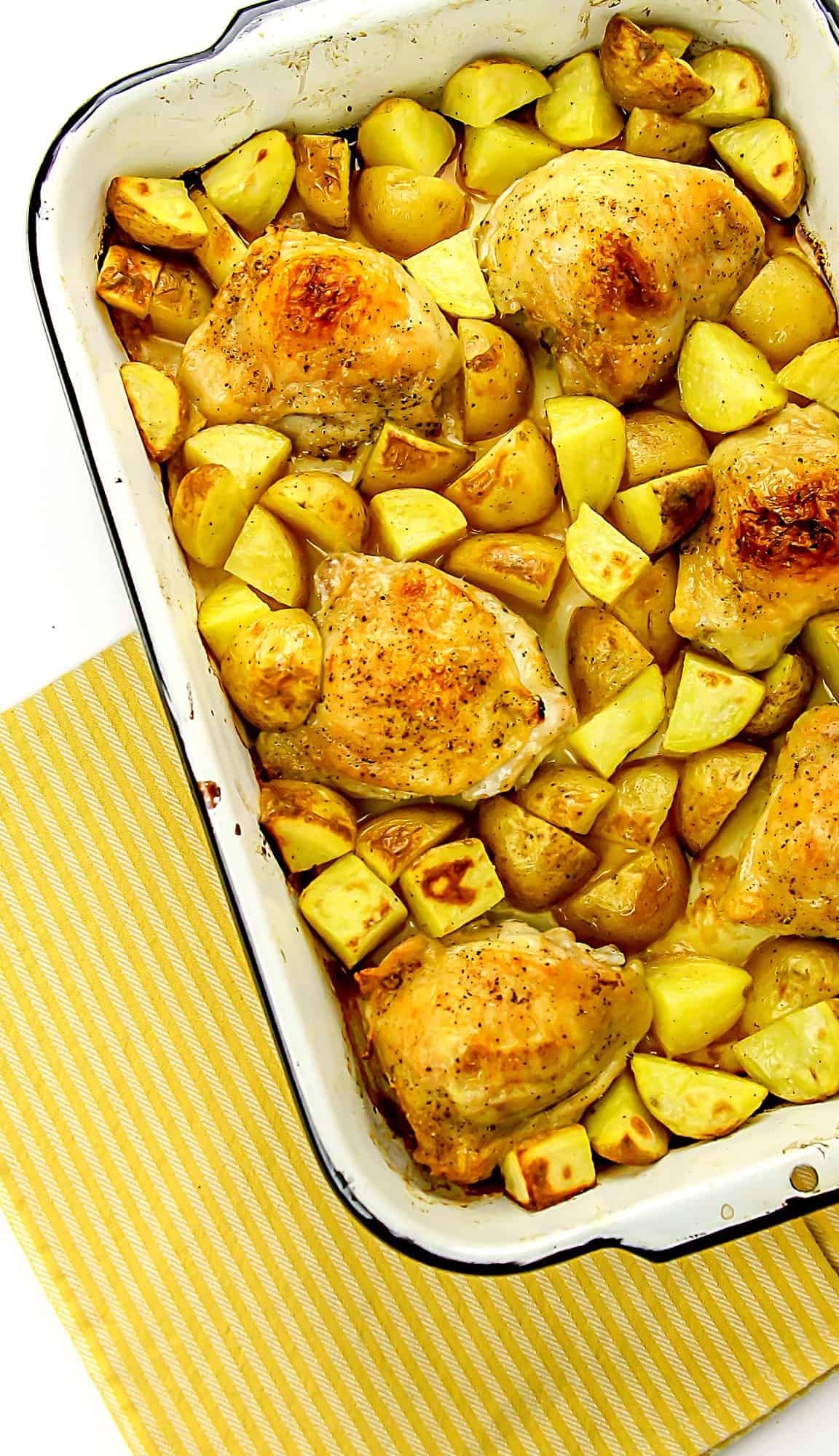 Baked chicken thighs with roasted potato chunks in a white rectangular baking dish on a yellow striped cloth.