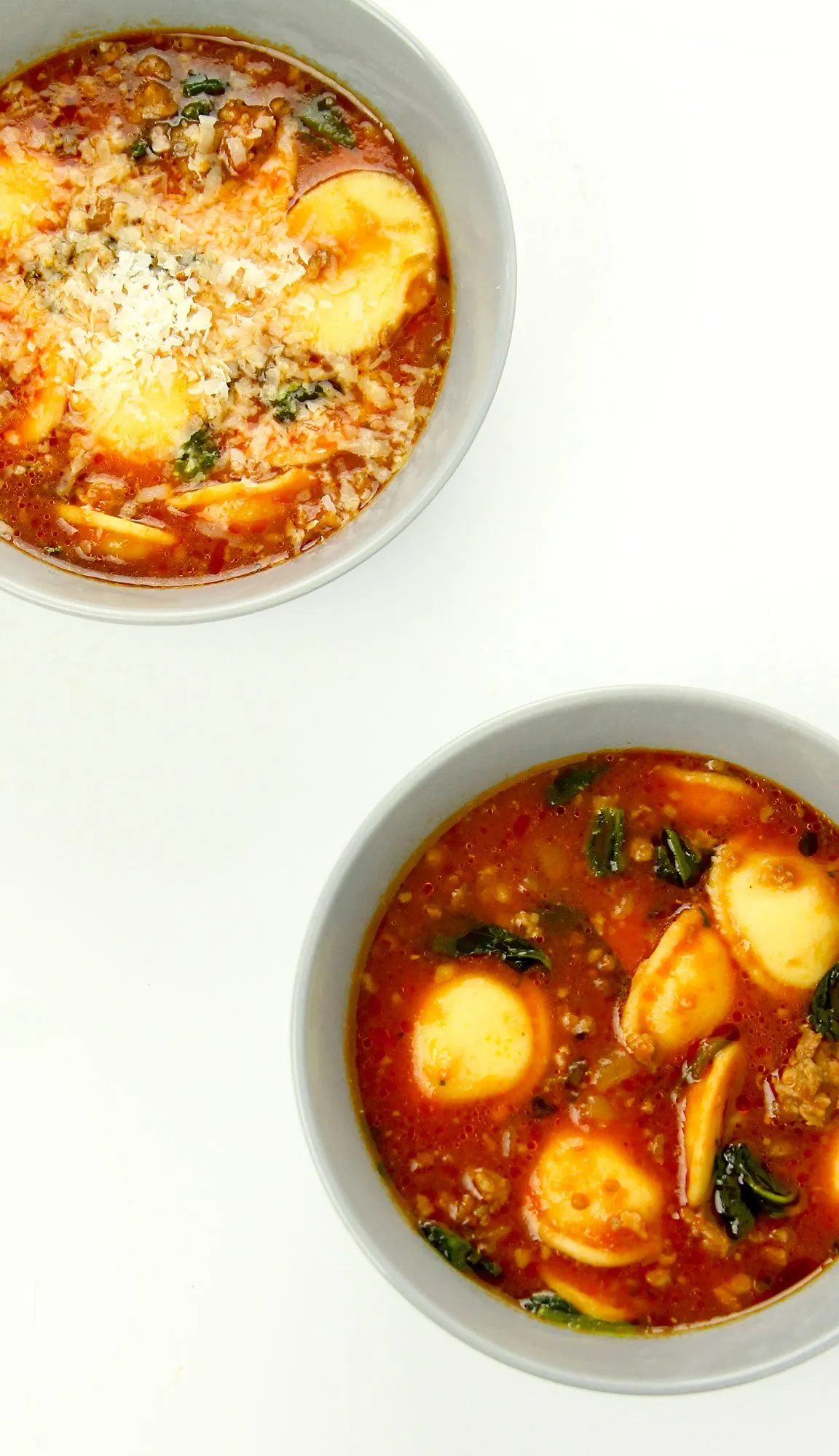 Two bowls of tomato-based soup with spinach, ground meat, and tortellini, one topped with grated cheese, on a white background.