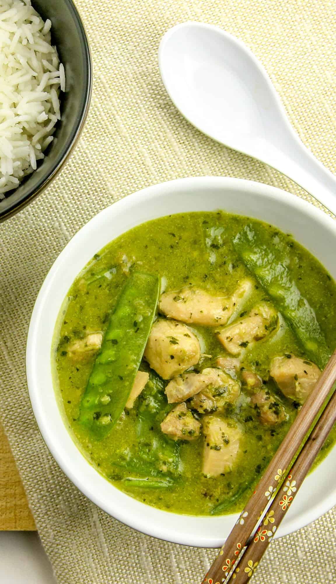 Chicken green curry with snow peas in a white bowl, wooden chopsticks, white rice in a black bowl, and a white spoon on a beige mat.