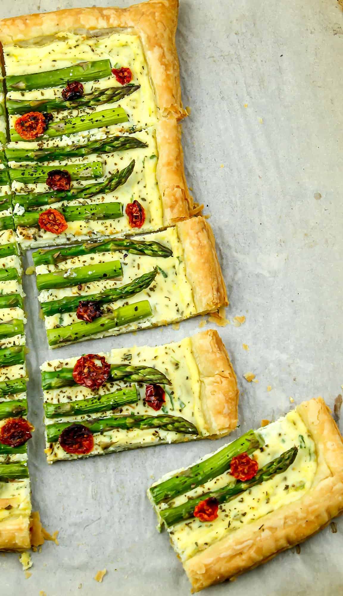 Asparagus tart with cherry tomatoes and herbs on a flaky pastry crust, cut into rectangular slices on a baking sheet.