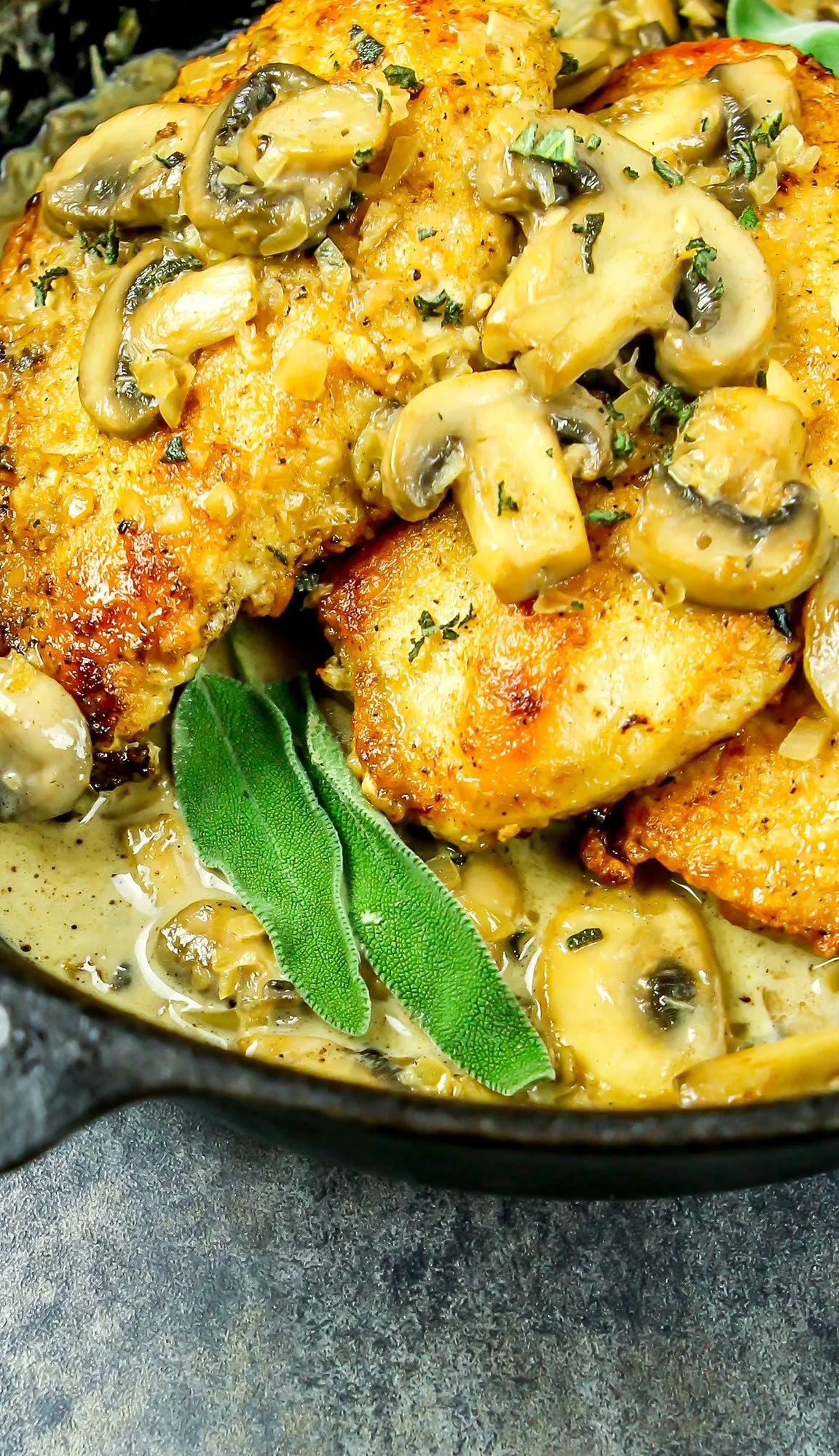 Pan-fried chicken breasts topped with sautéed mushrooms and fresh sage leaves in a creamy sauce in a cast iron skillet.