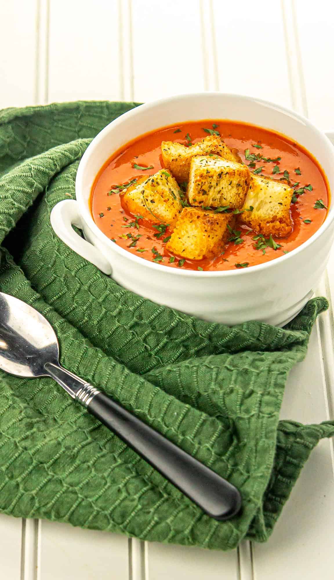 White bowl of tomato soup topped with herbed croutons and parsley, placed on a green textured cloth with a spoon beside it.