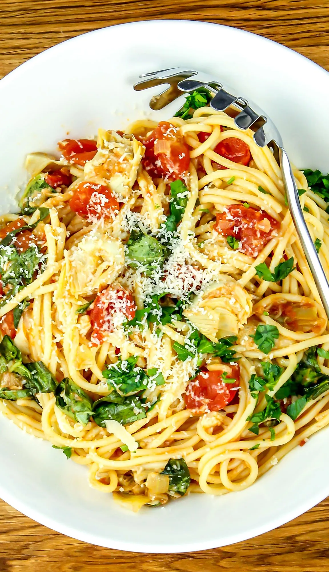 Spaghetti pasta with cherry tomatoes, spinach, artichoke hearts, grated cheese, and parsley in a white bowl with a pasta fork.