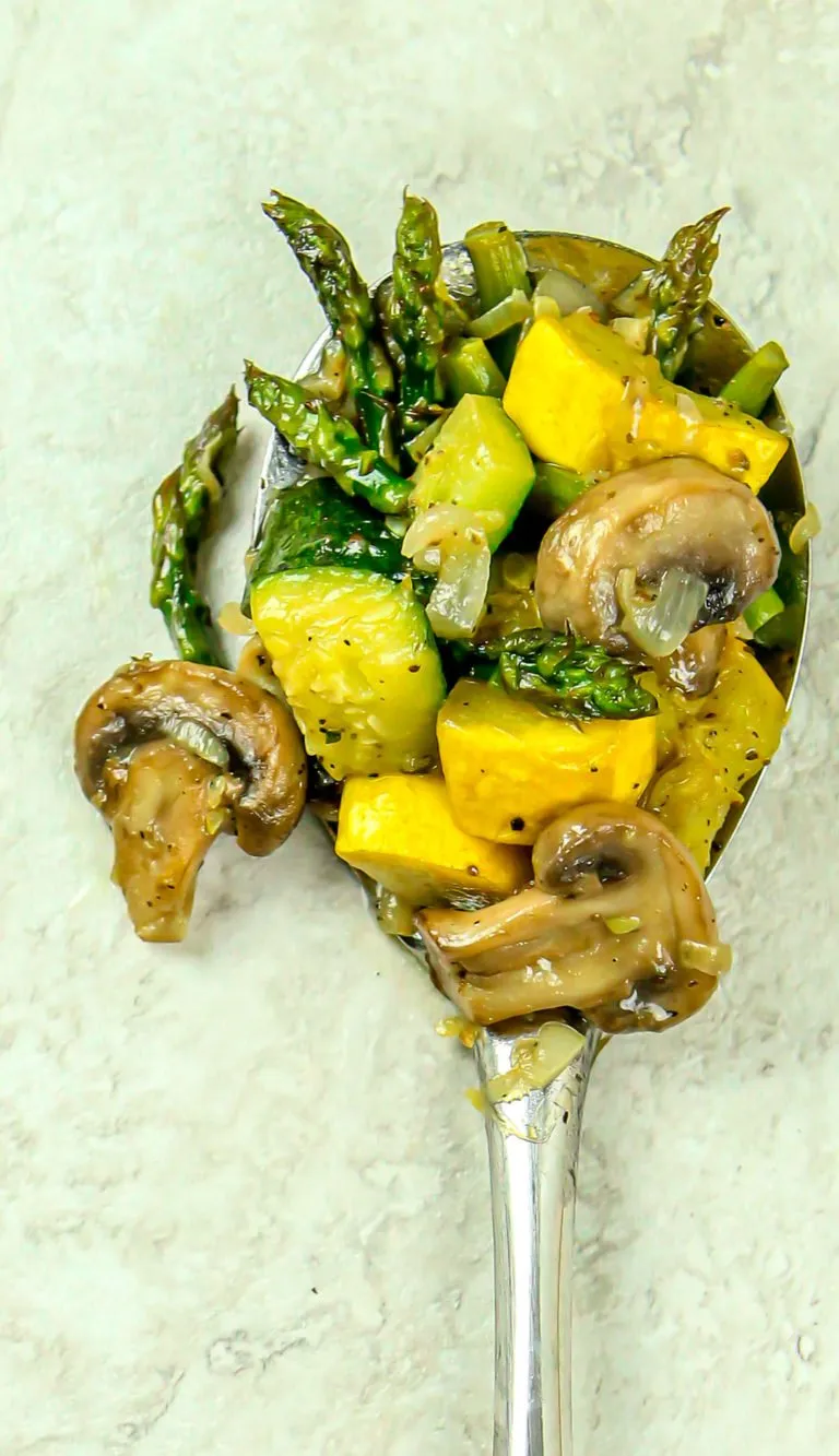 Sautéed mixed vegetables including asparagus, yellow squash, zucchini, mushrooms, and onions on a silver spoon.