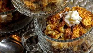 Glass cups filled with toasted bread pudding topped with a dollop of whipped cream on a textured surface with spoons nearby.