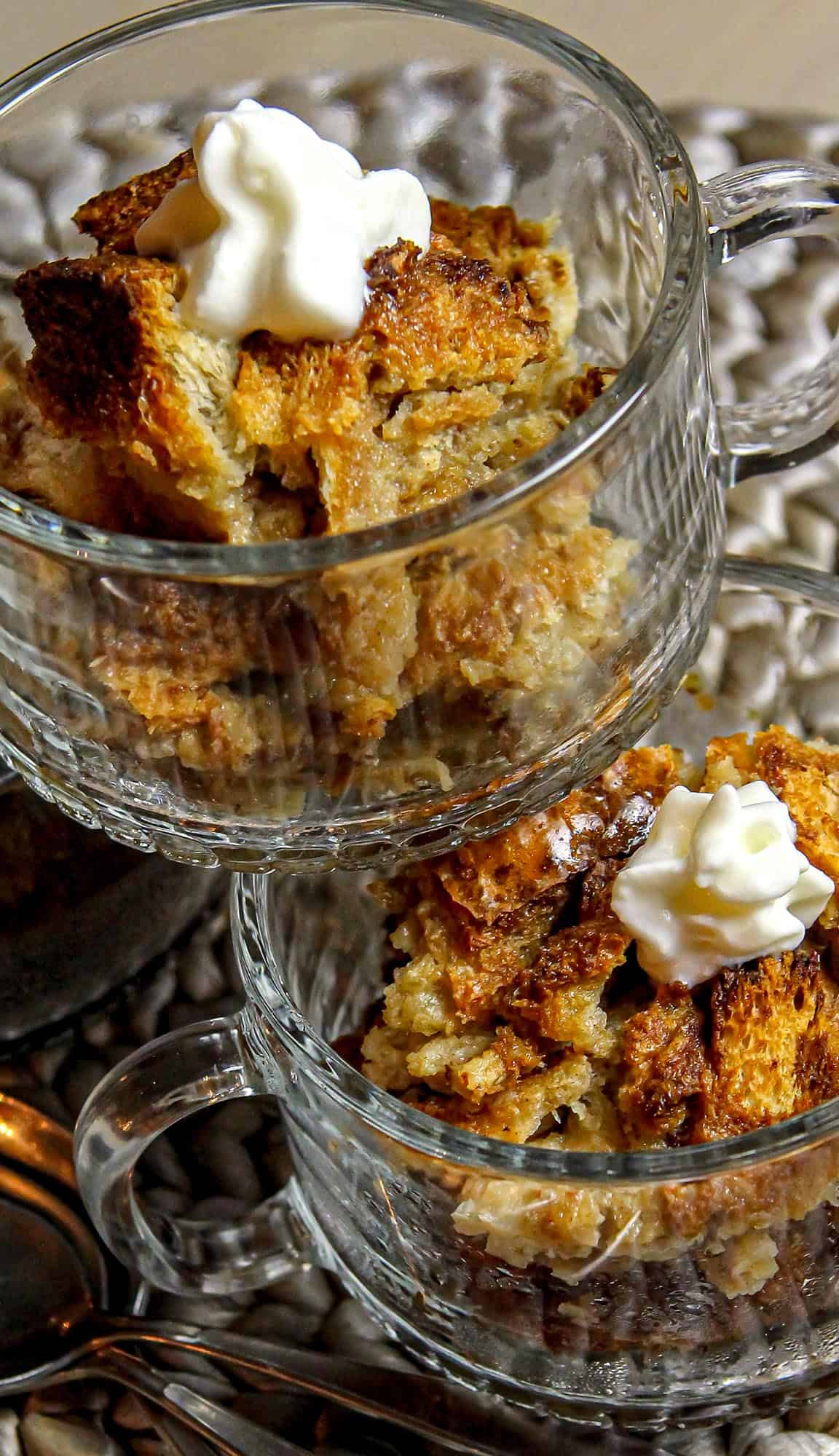 Two glass cups filled with toasted bread pudding topped with whipped cream on a textured beige surface.